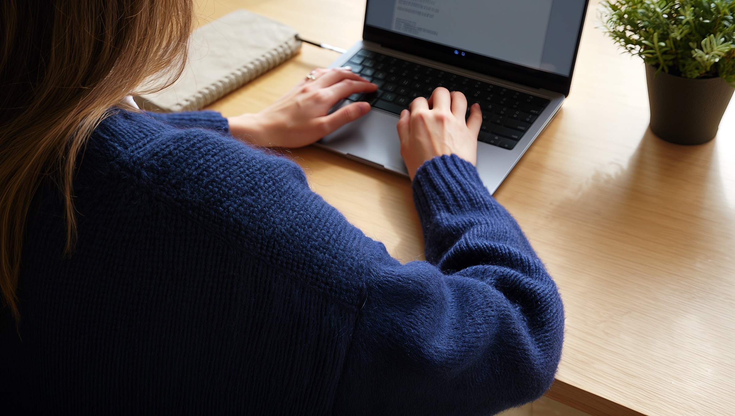A person in a blue sweater typing on a laptop at a wooden desk.